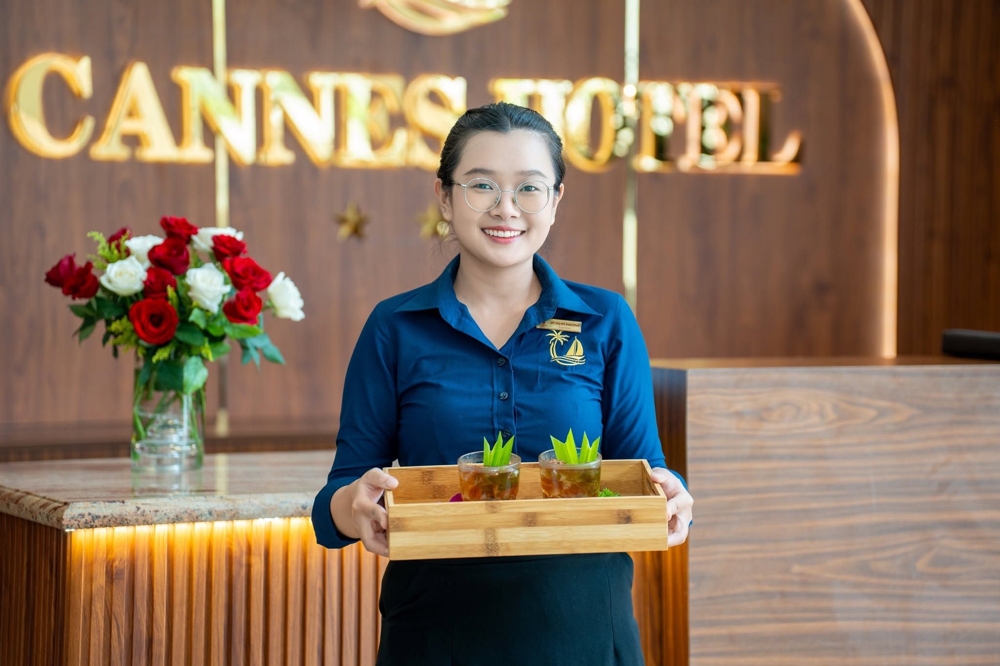 A hotel receptionist holding a tray with two drinks and a floral arrangement.