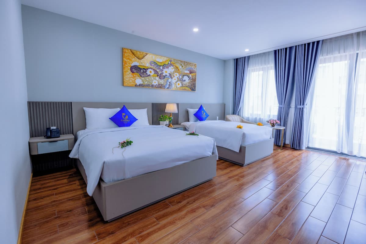A hotel room with two beds, a large painting on the wall, and a view of the outdoors through sheer curtains.