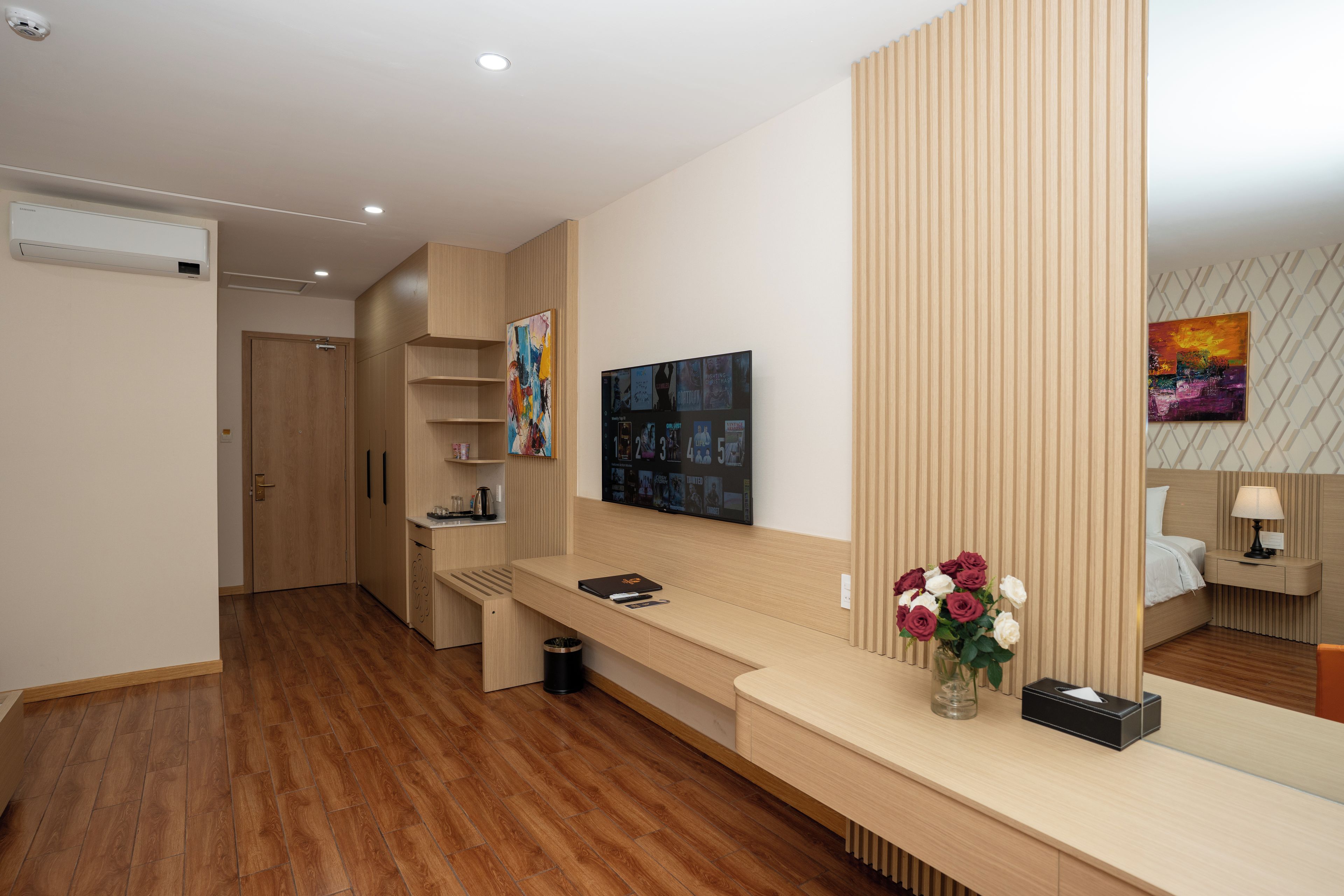 A modern hotel room with a wooden floor, a reception desk, a large abstract painting, and a bed with a nightstand.