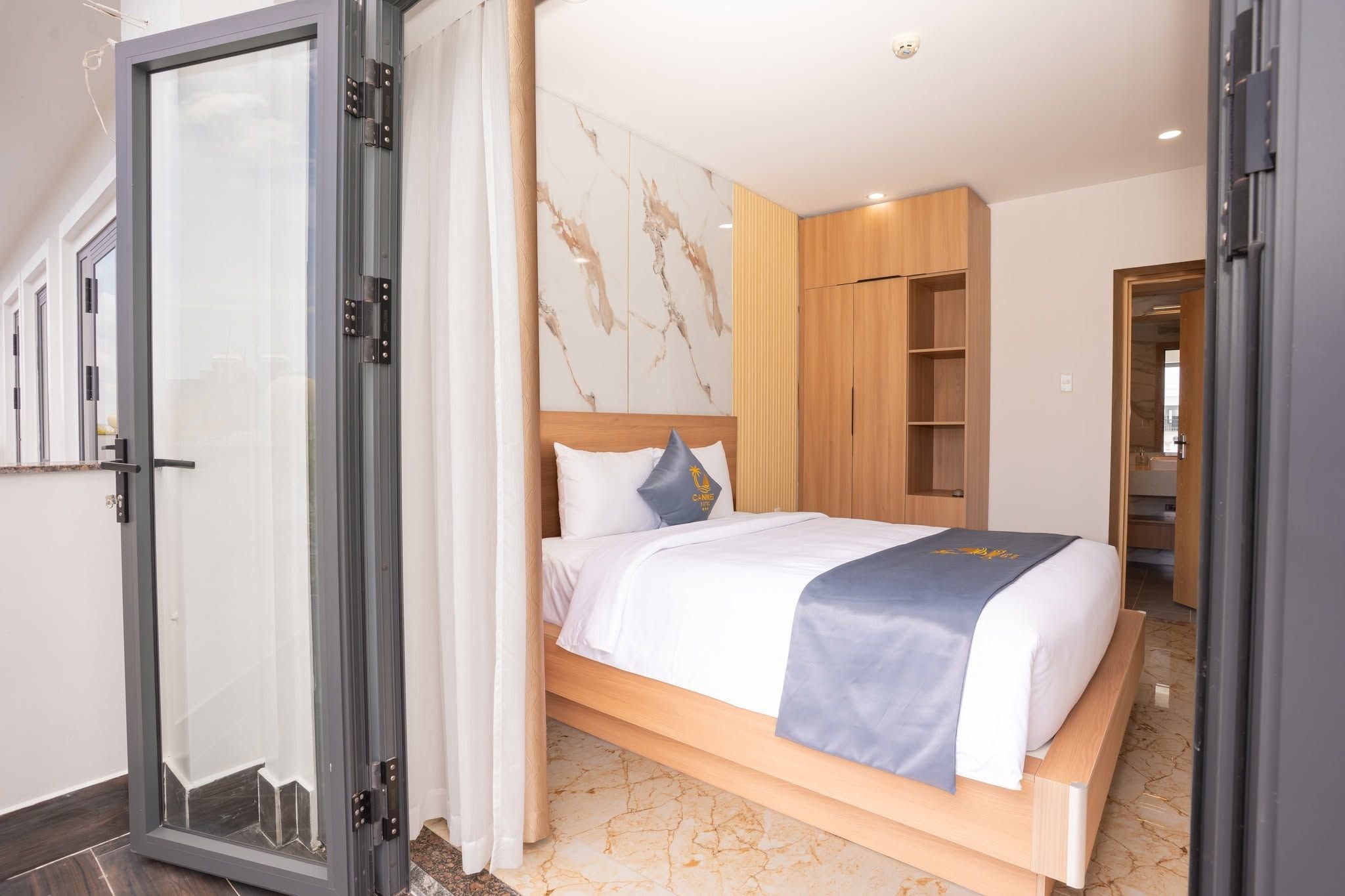 Une chambre d’hôtel moderne, équipée d’un lit, d’une armoire en bois et d’une porte vitrée donnant sur un balcon.