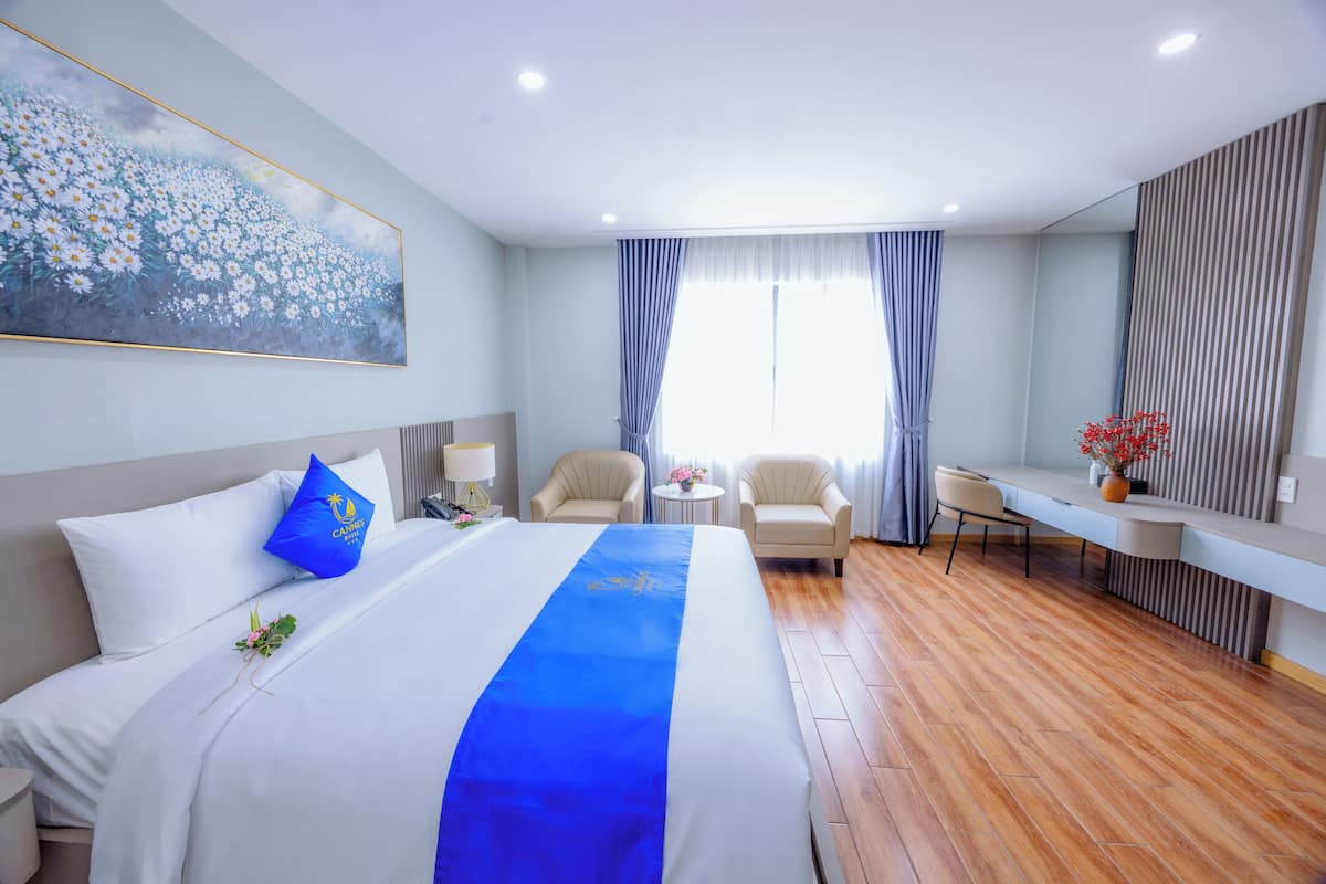 A hotel room with a large bed, two armchairs, a desk, and a large window with curtains.