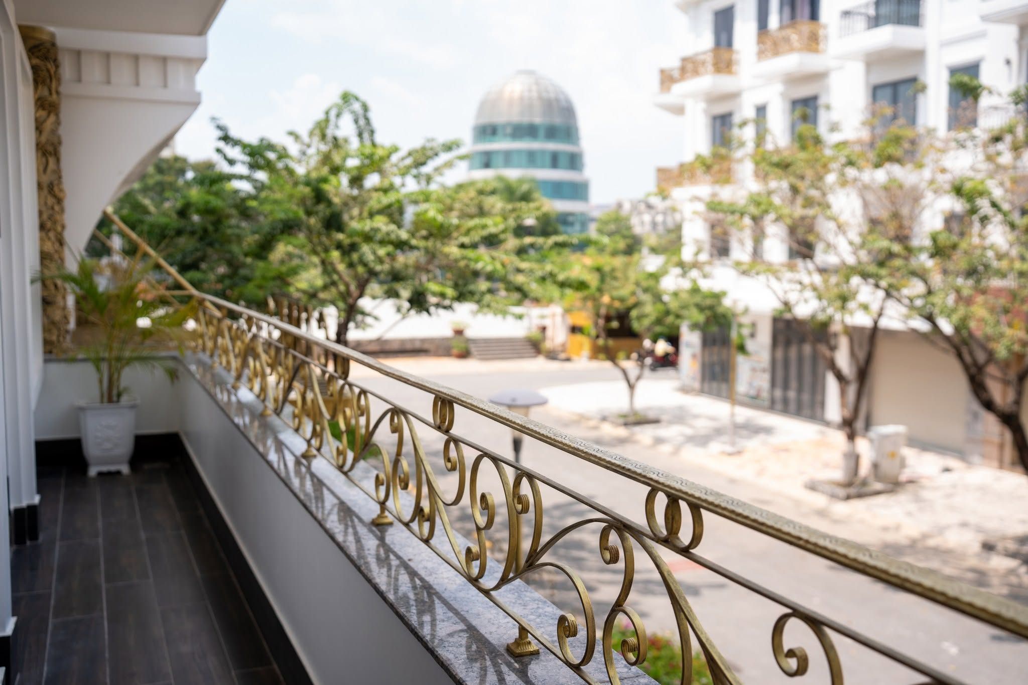 A balcony with ornate metal railings overlooking a street with buildings and trees.