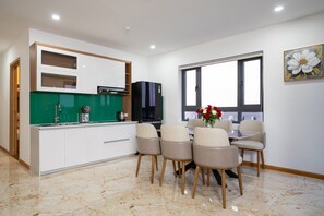 A modern kitchen with white cabinets, a green backsplash, and a black refrigerator. There is a dining area with beige chairs and a table, and a window with a view. - Cannes Hotel (Phu Khuong)