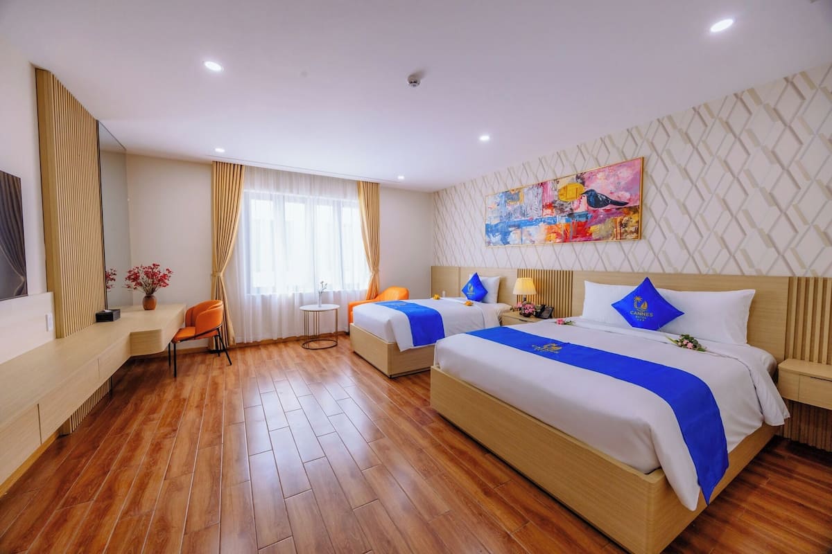 A hotel room with two beds, a wooden floor, a desk with a chair, a small table, and a painting on the wall.