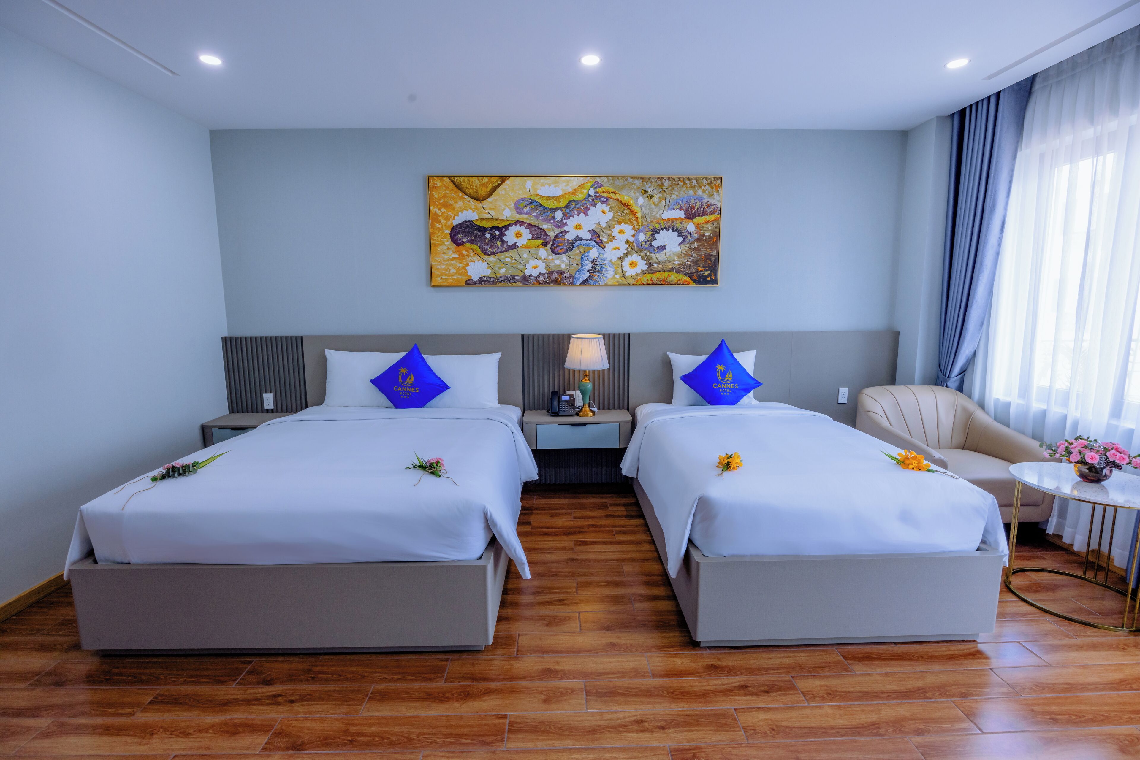 A hotel room with two beds, a nightstand, a lamp, a chair, and a painting on the wall.
