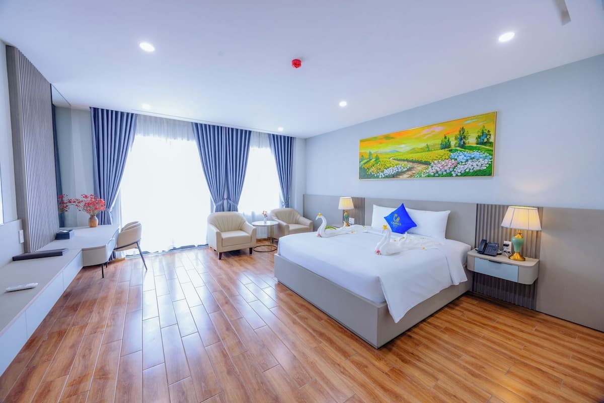 A hotel room with a large bed, two bedside tables, a desk, and a chair. There is a painting on the wall and a window with curtains.