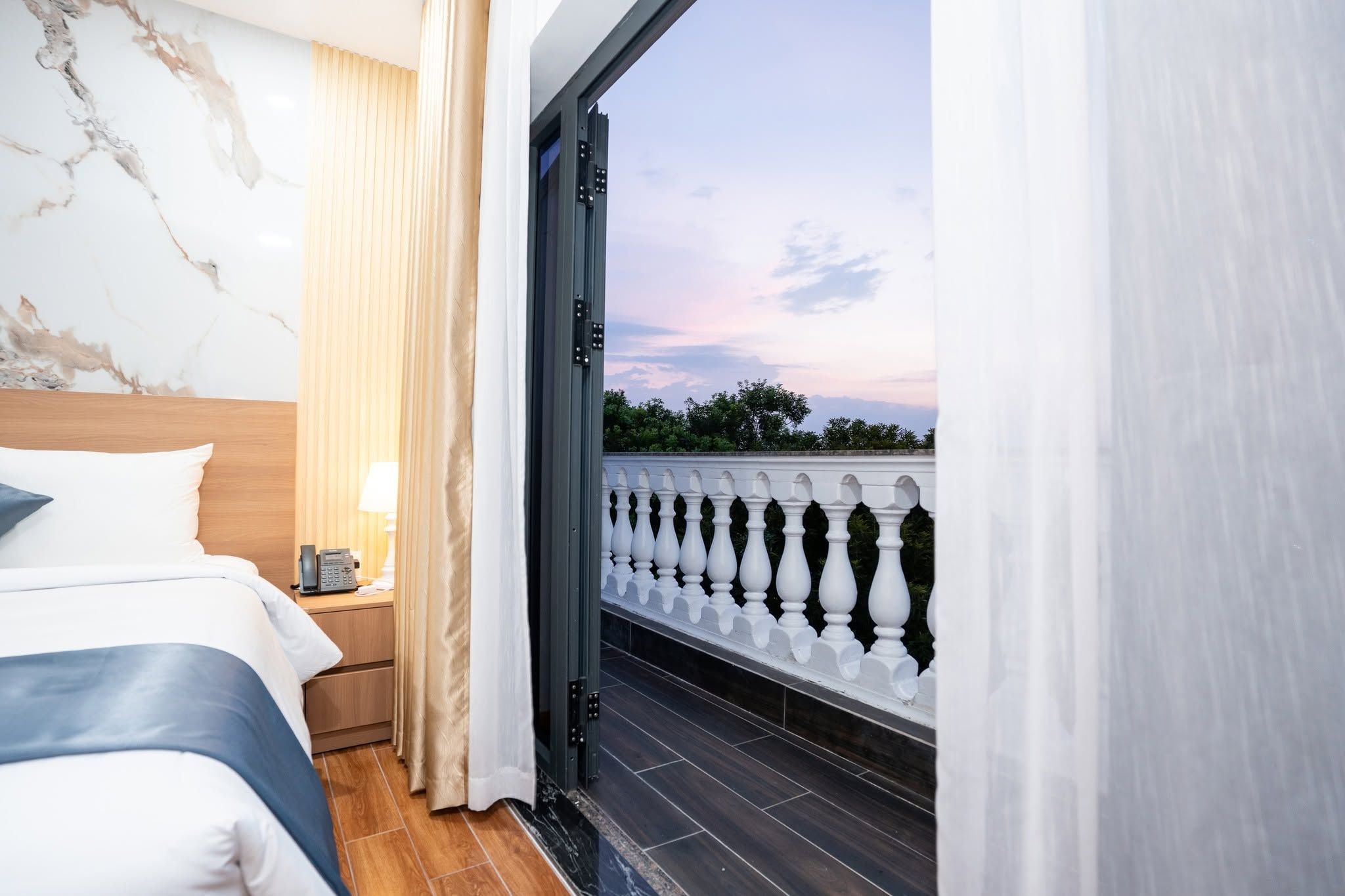 A hotel room with a bed, bedside table, lamp, and a view of a balcony with a balustrade.