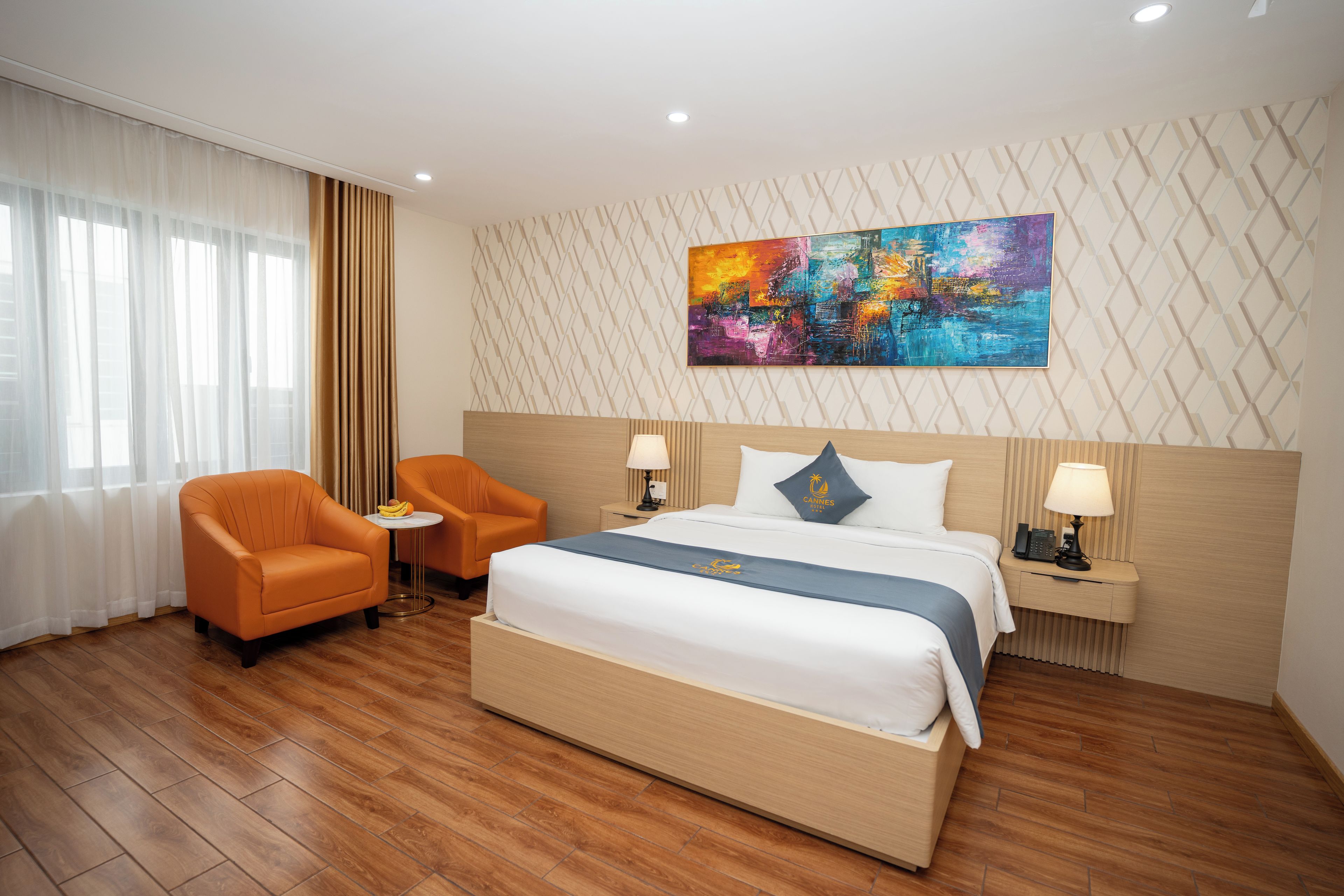 A hotel room with a large bed, two armchairs, a small table, a nightstand with a lamp, and a painting on the wall.