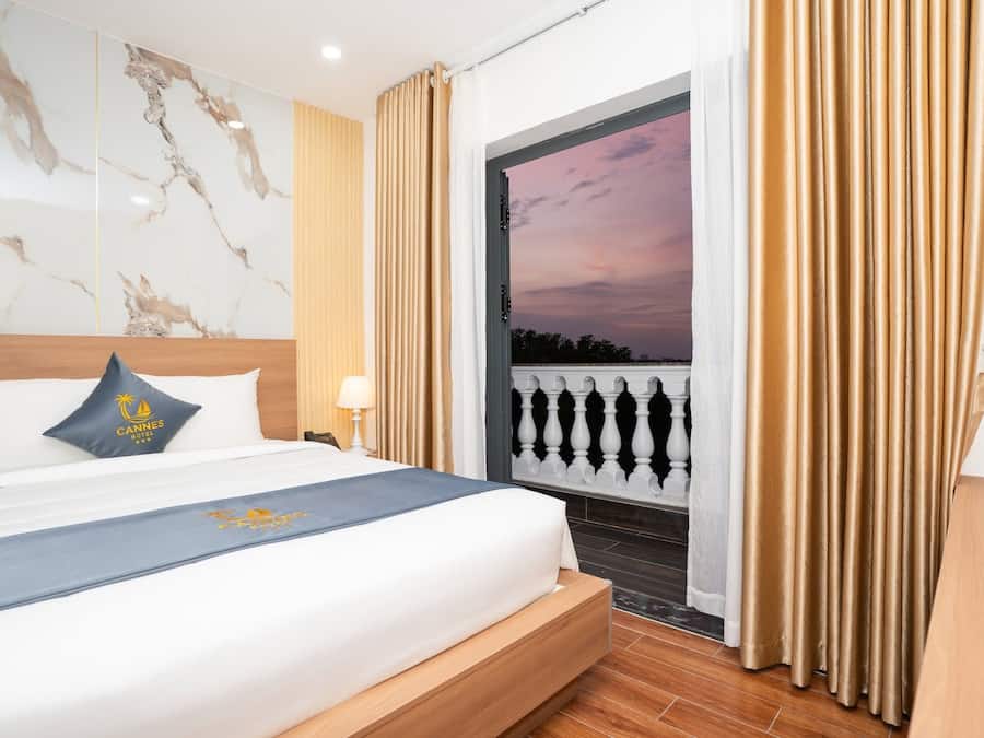 Una habitación de hotel con una cama, una mesita de noche, una lámpara, una ventana con cortinas y vistas al cielo.