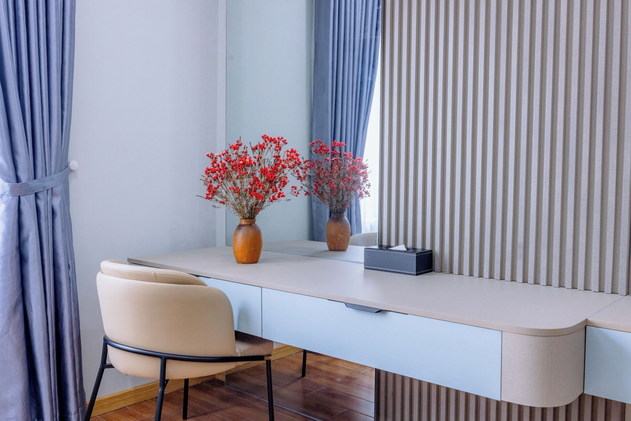 Een modern bureau met een stoel, twee vazen met rode bloemen, een spiegel en een gestreept gordijn.