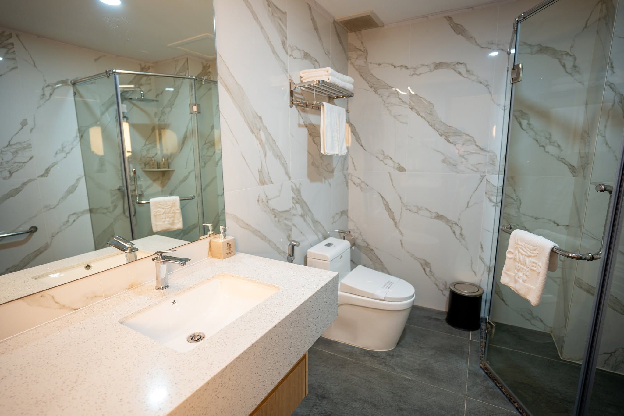 Un baño moderno con detalles de mármol, cabina de ducha con mampara de vidrio, inodoro y lavamanos con espejo.