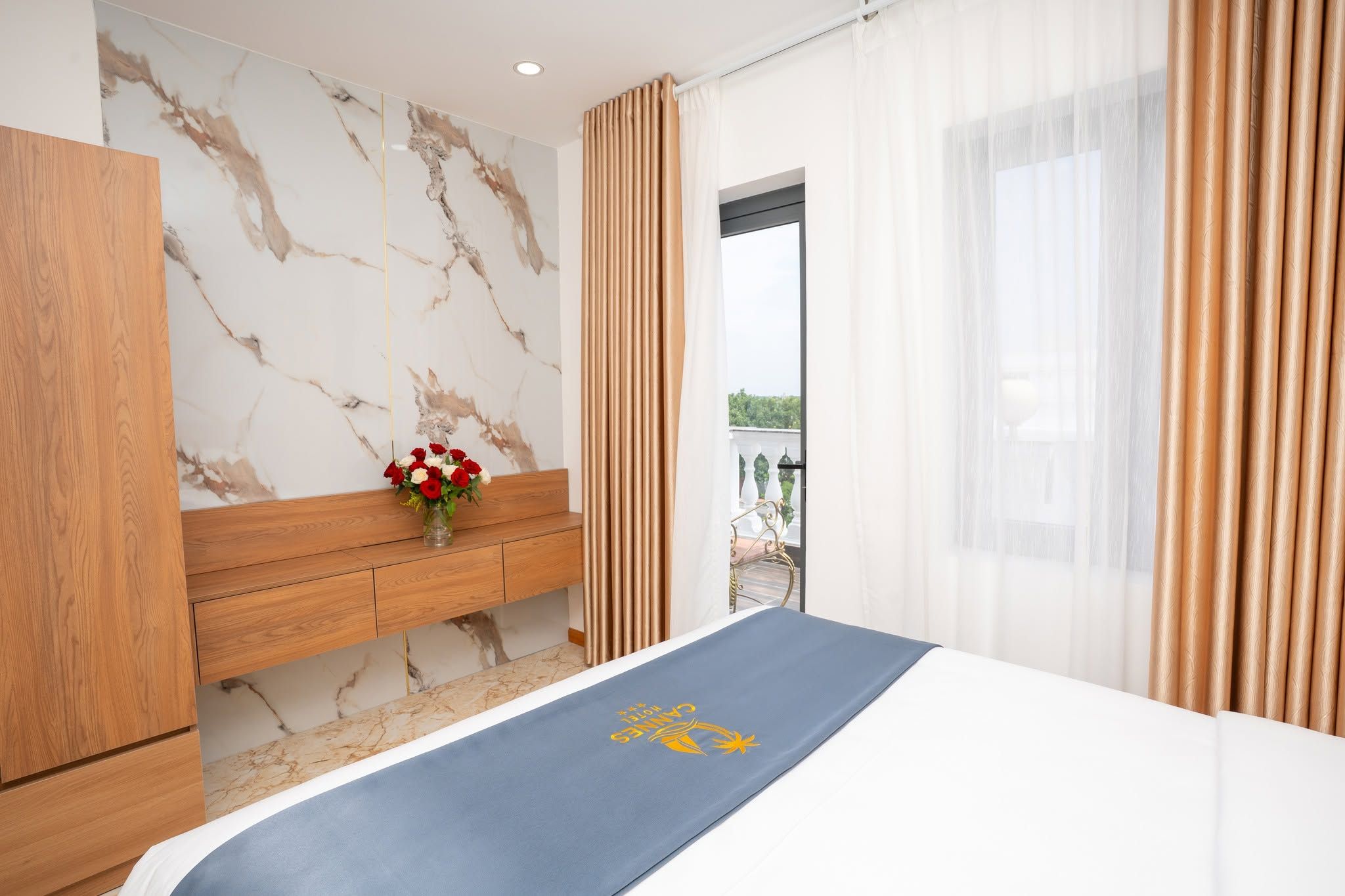 A hotel room with a marble wall, a wooden bench, a bed with a blue bedspread, and a window with curtains.