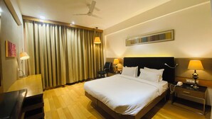 A hotel room with a large bed, a desk, a chair, a nightstand with a lamp, a ceiling fan, and a window with curtains. - The Manak Bagh (Sehore)