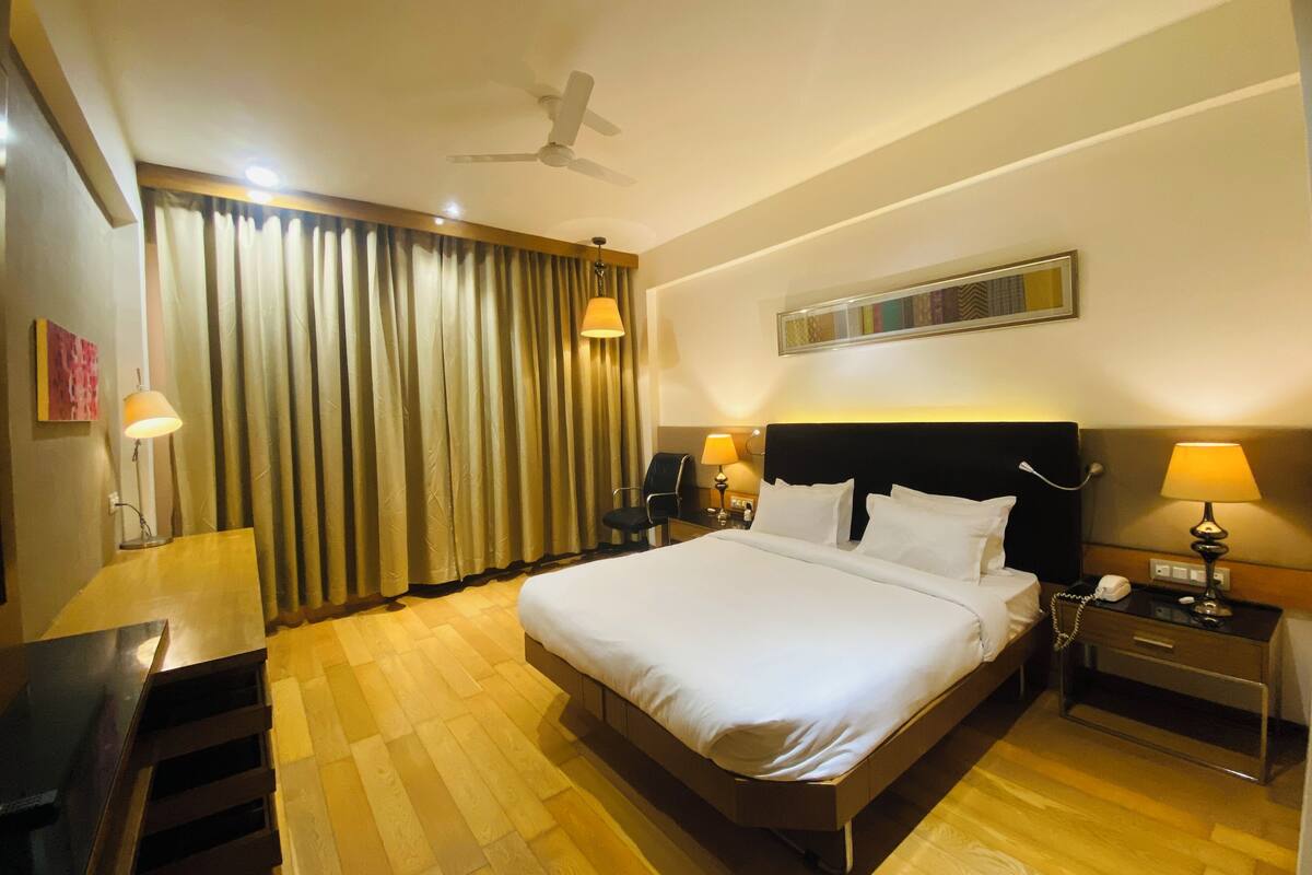 A hotel room with a large bed, a desk, a chair, a nightstand with a lamp, a ceiling fan, and a window with curtains.
