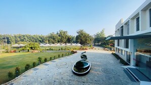 A courtyard with a paved walkway, a modern building with large windows, and a grassy area with trees. - The Manak Bagh (Sehore)