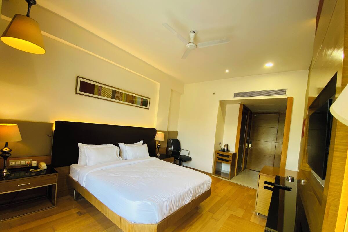 A hotel room with a large bed, bedside tables, a desk, a chair, a TV, a ceiling fan, and a door leading to another room.