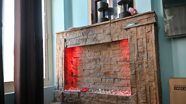 Una chimenea de piedra con interior iluminado en rojo, un estante de piedra con adornos, y una ventana con cortinas.