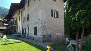 A rustic two-story building with a sloped roof, a balcony, and a small garden area with grass and flowers.