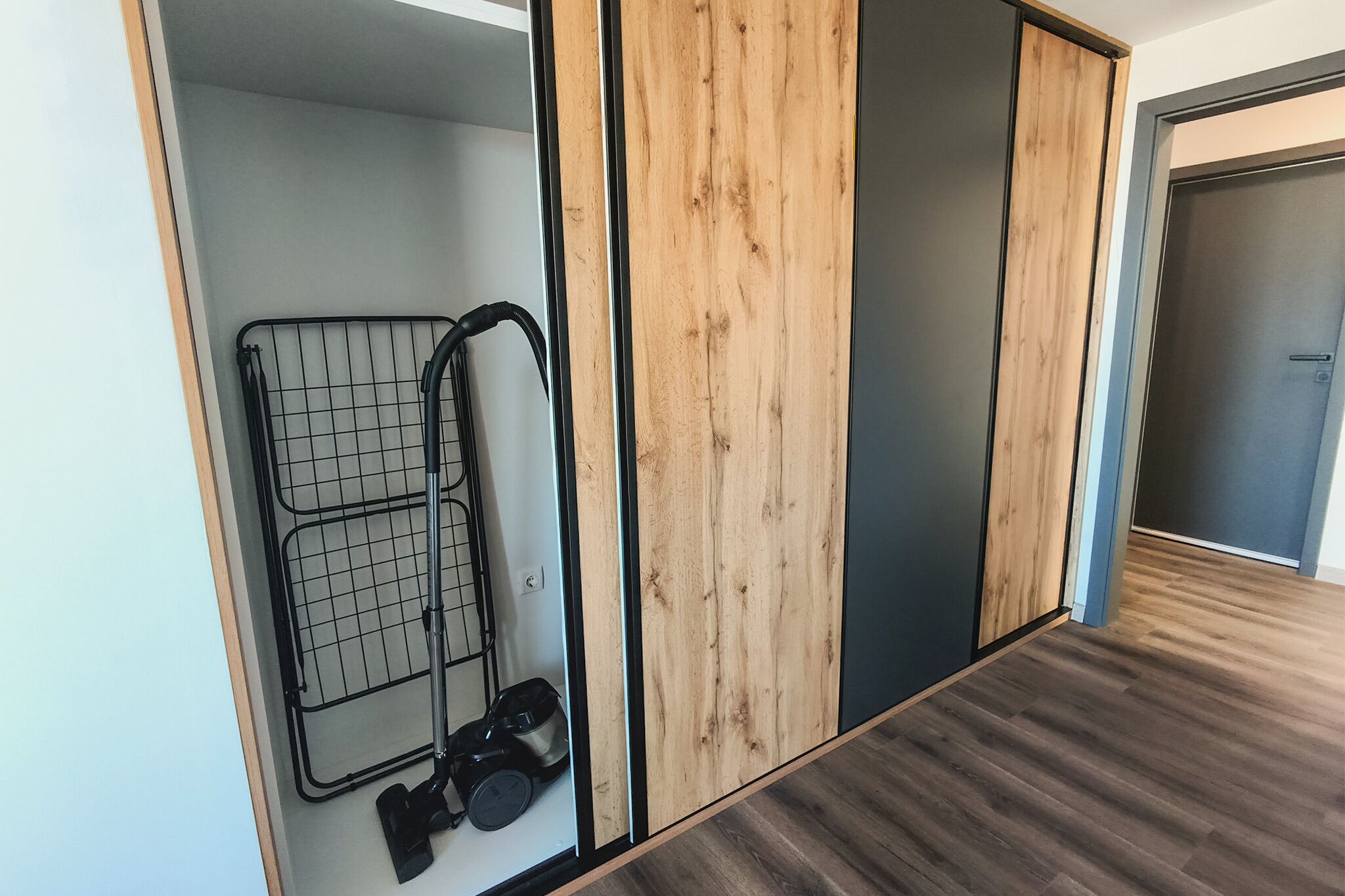 Une pièce avec un plancher en bois, un étagère en métal sur laquelle se trouve un aspirateur, et une porte avec un panneau en bois.