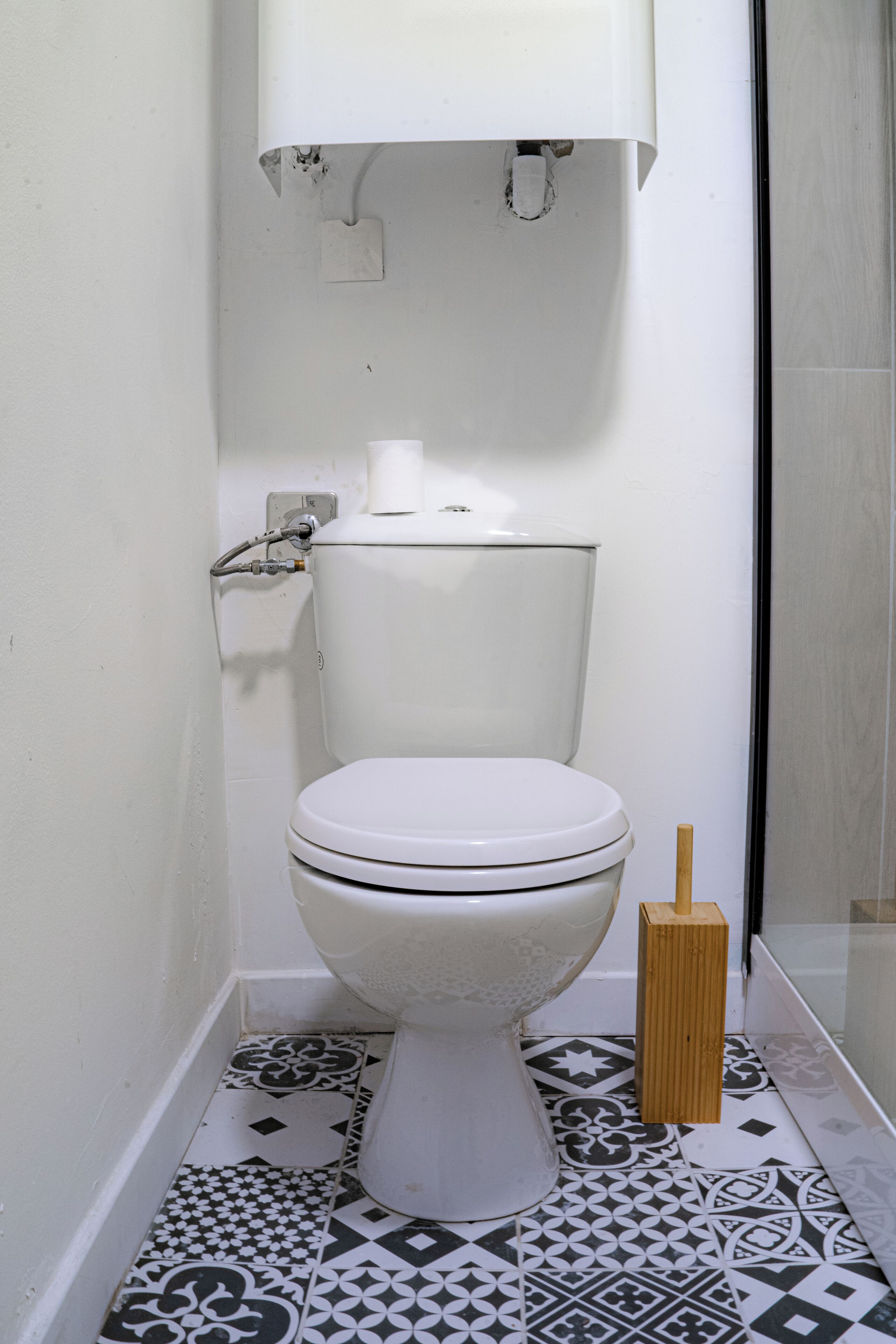 A toilet with a white tank and lid, a toilet paper holder, and a bidet sprayer. The floor has patterned tiles.