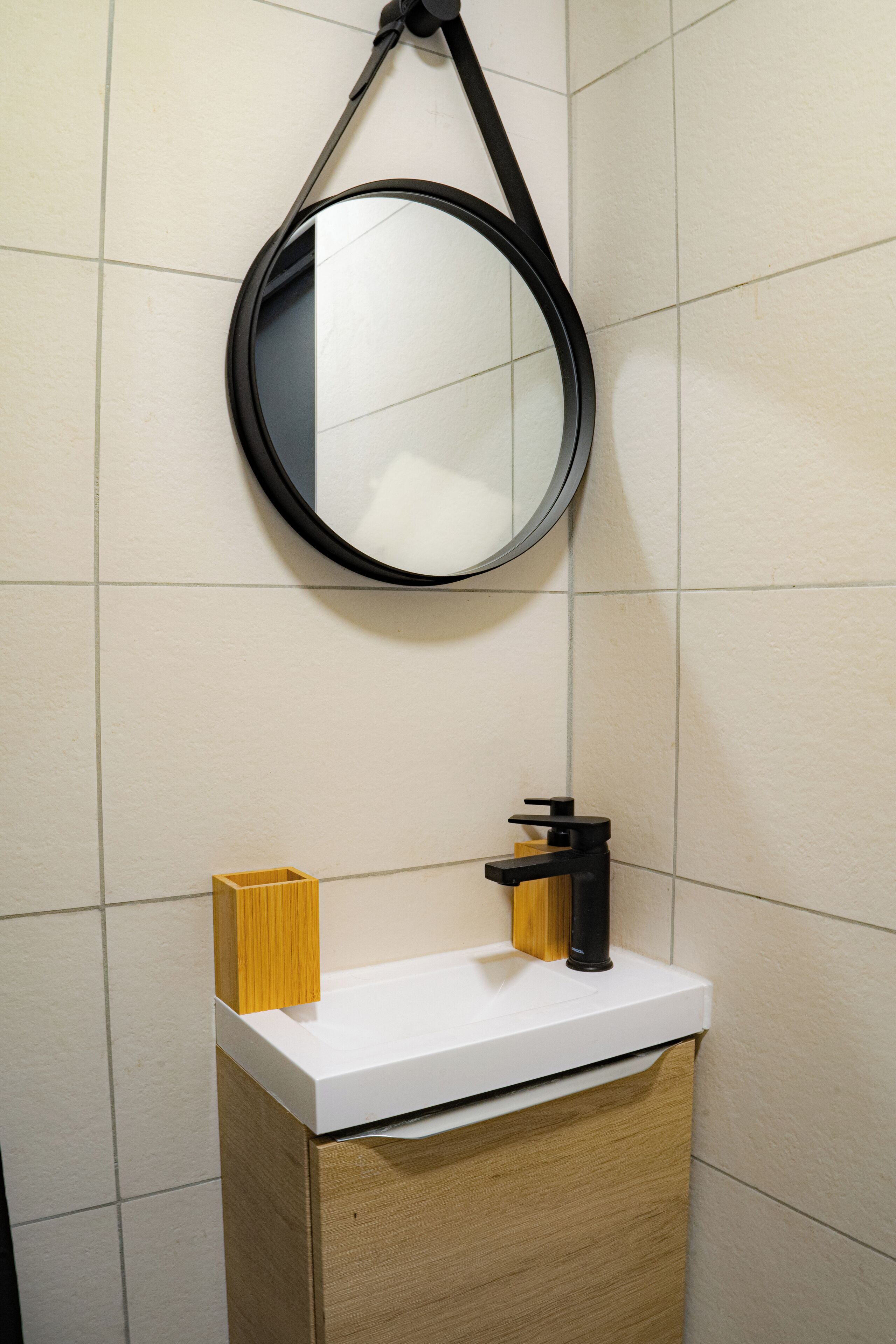 Een badkamer met een wit aanrechtblad, een houten kastje, een zwarte kraan en een ronde spiegel met een zwarte lijst.