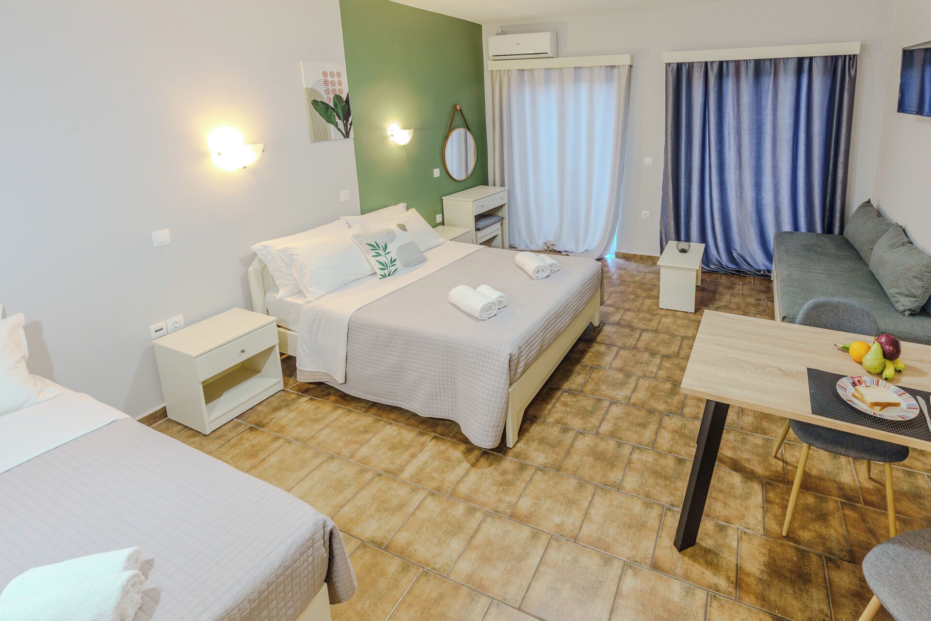Habitación de hotel con cama, mesitas de noche, un escritorio con silla, una pequeña mesa con frutas, un espejo y una ventana con cortinas.