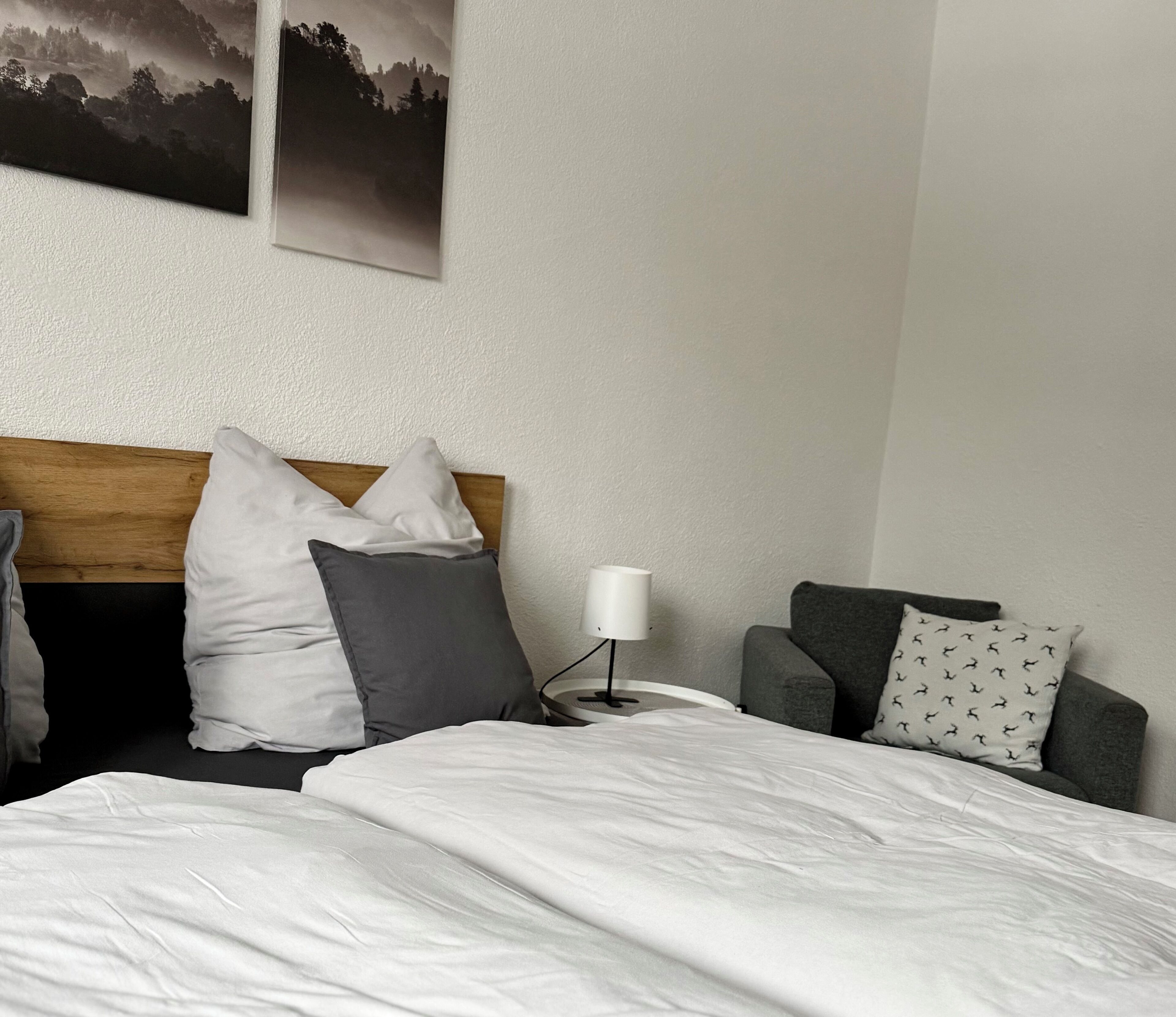 Una habitación de hotel con una cama, cabecera de madera, mesita de noche con lámpara, un sillón gris y un cuadro en la pared.