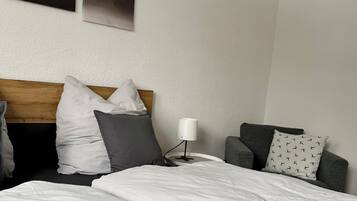 Una habitación de hotel con una cama, cabecera de madera, mesita de noche con lámpara, un sillón gris y un cuadro en la pared.
