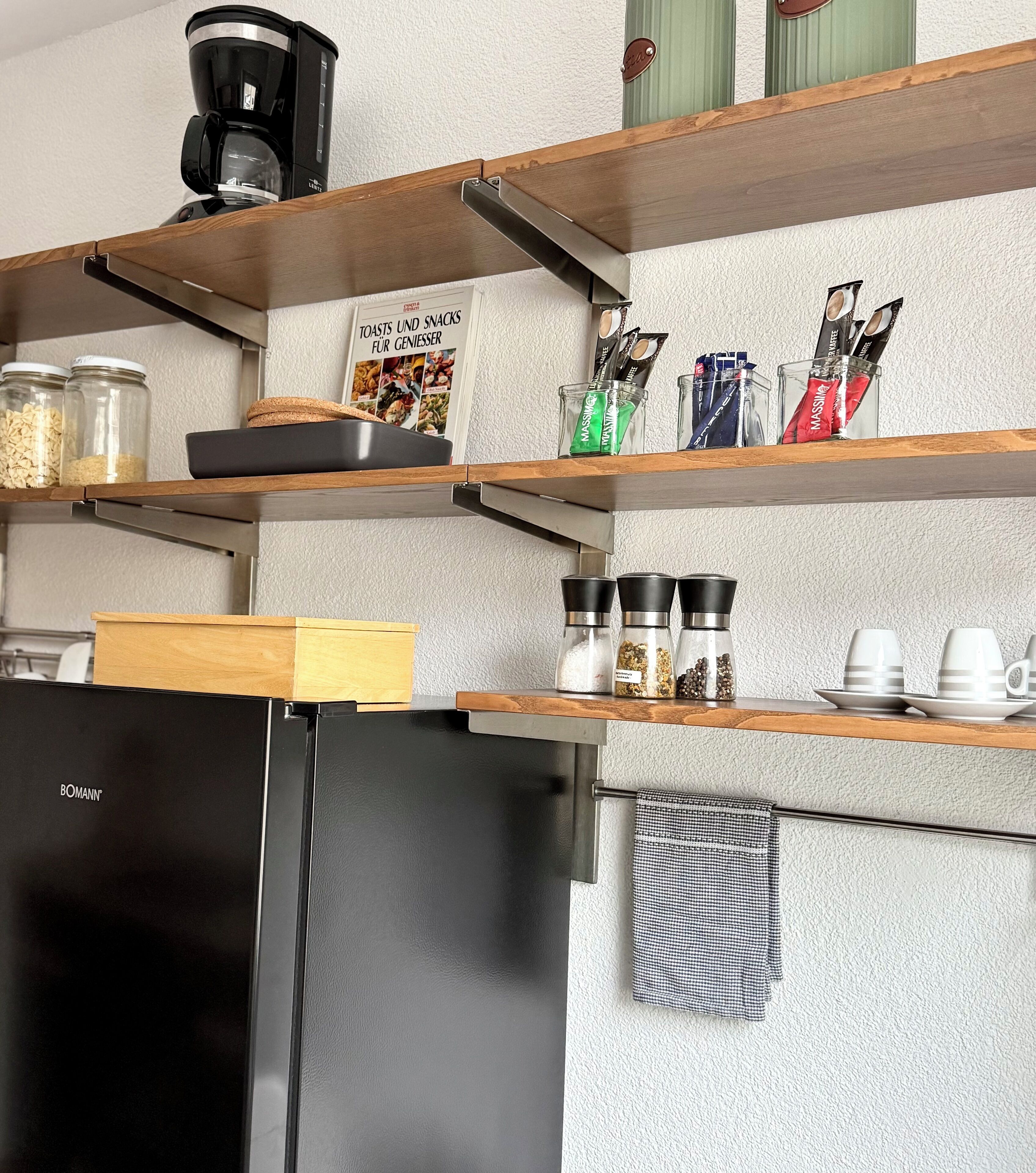 Una cocina con estantes de madera, un refrigerador negro y diversos utensilios de cocina.