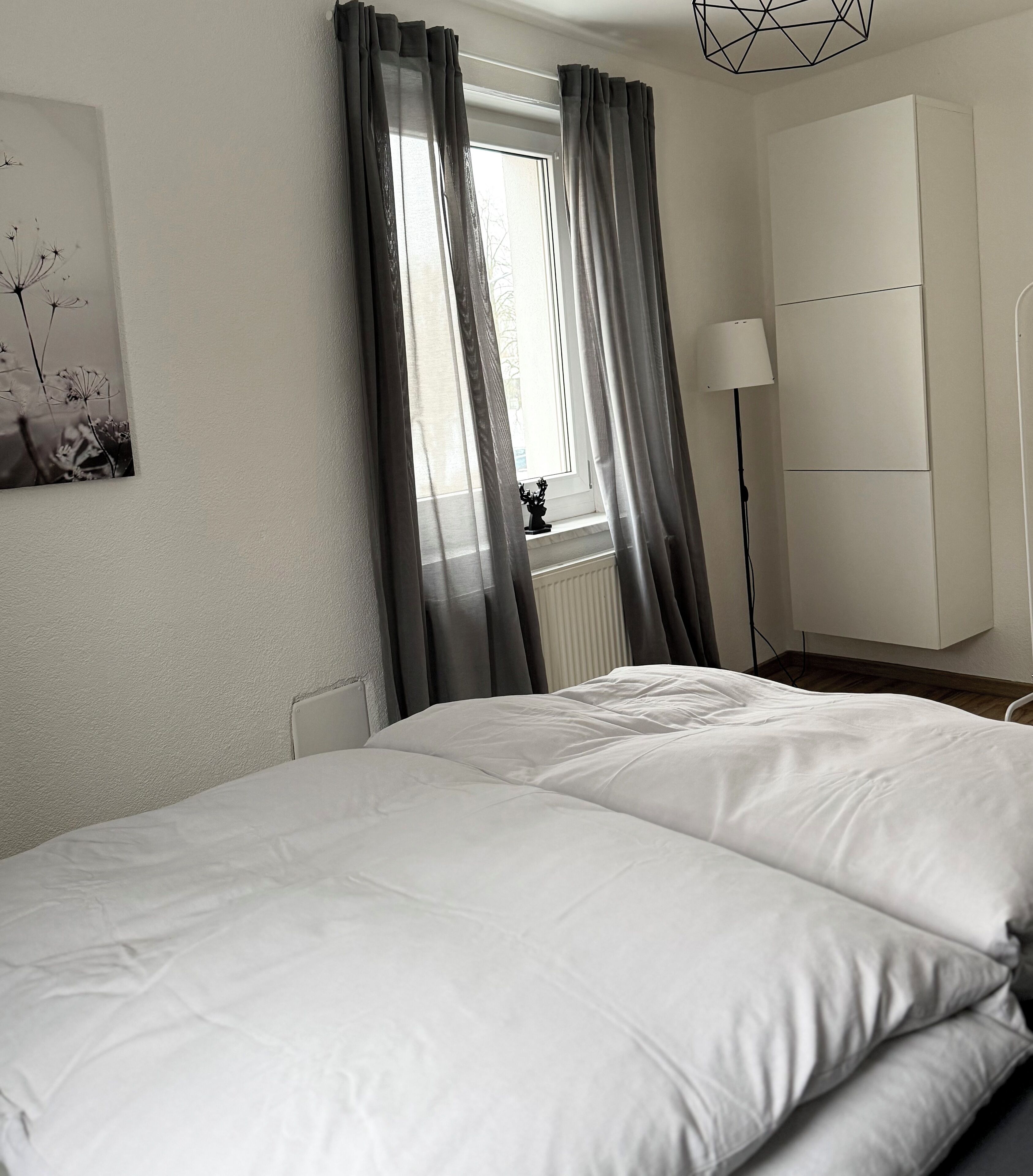 A neatly made bed with white bedding, a window with grey curtains, a floor lamp, and a white wardrobe.