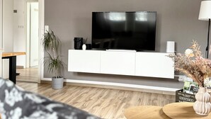 Uma sala de estar moderna com uma televisão de tela plana sobre um armário branco, uma planta em vaso, uma pequena mesa com um vaso e um sofá estampado.