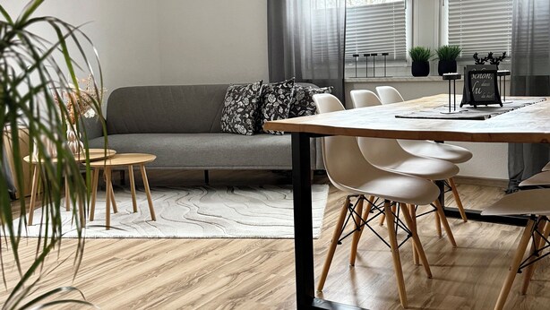 Uma sala de estar moderna com um sofá cinza, uma mesa de jantar de madeira e um tapete branco.