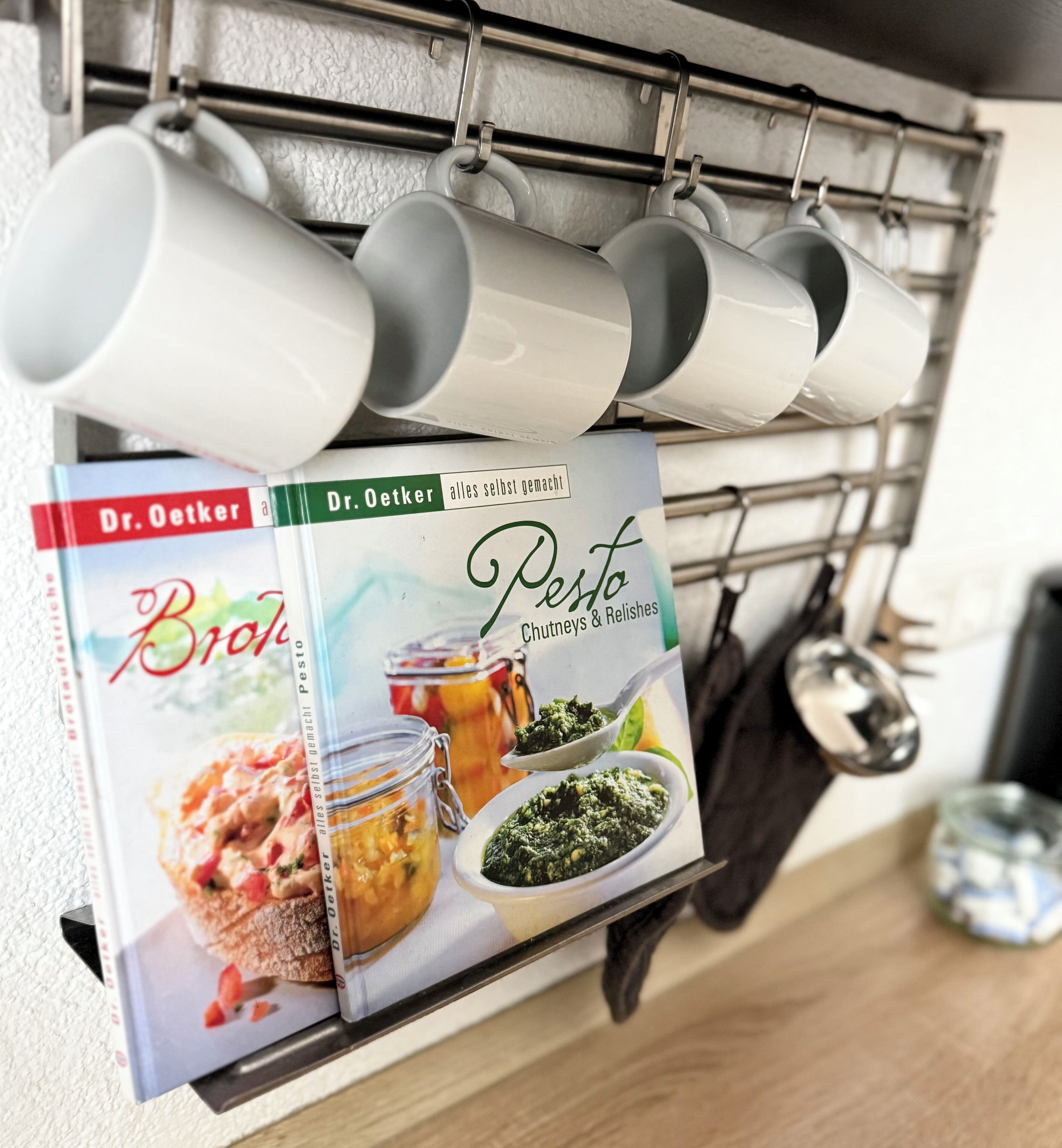 Eine Küche mit einer Wandablage, auf der mehrere weiße Tassen hängen. Darunter liegt ein Kochbuch mit dem Titel 'Dr. Oetker Pesto' und Abbildungen von Pesto sowie anderen Gerichten.