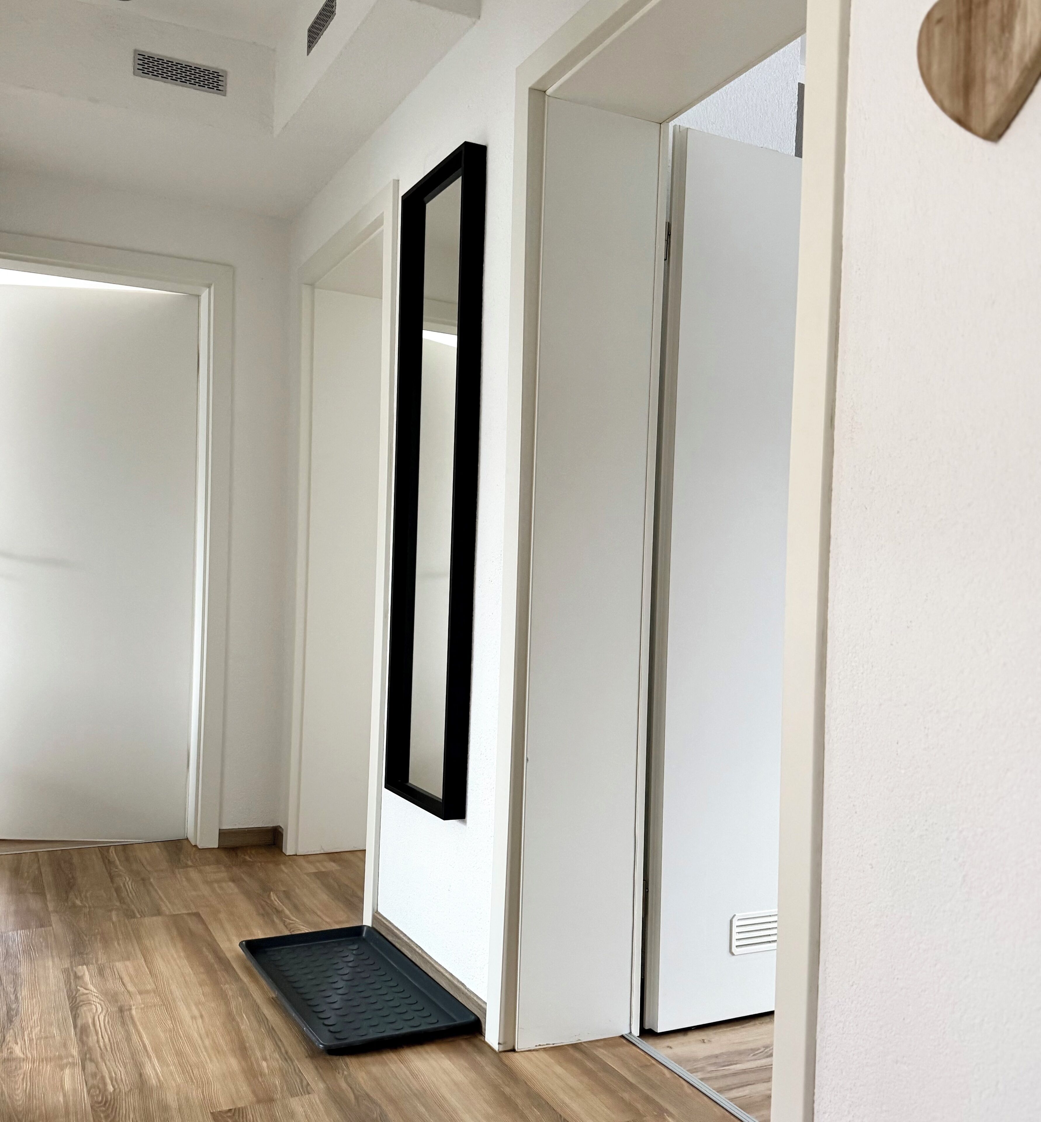 A hallway with a wooden floor, white walls, and a black-framed mirror on the wall.