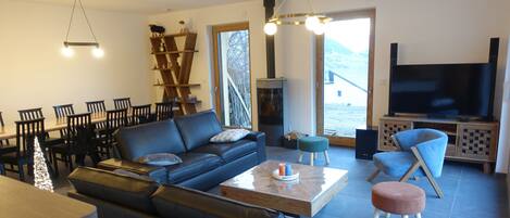 A modern living room with a sofa, coffee table, and TV. There is a fireplace, dining area, and a view of the outdoors.