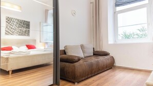 A modern living room with a brown leather sofa, a wooden floor, a large window with curtains, and a visible clock on the wall. - Bright 1BR Apt Perfect for Long Stays (Vienna)