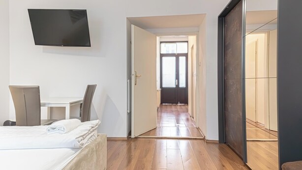A hotel room with a bed, a desk, a chair, a TV mounted on the wall, and a view through an open door to a hallway. - Bright 1BR Apt Perfect for Long Stays (Vienna)