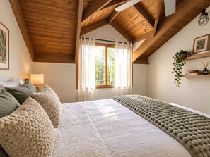 Quarto com teto de madeira, uma cama grande com manta texturizada, mesa de cabeceira com abajur, janela com cortinas leves e uma estante com planta e livros.