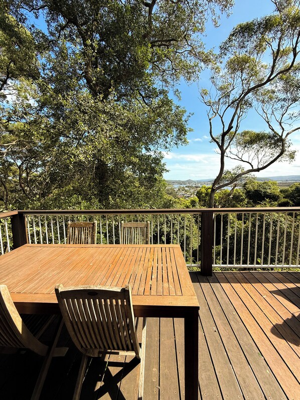 A wooden deck with a table and chairs, overlooking a scenic landscape with trees and a distant view of a town.