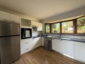 A modern kitchen with white cabinets, a stainless steel refrigerator, a dishwasher, and a window with a view of greenery.