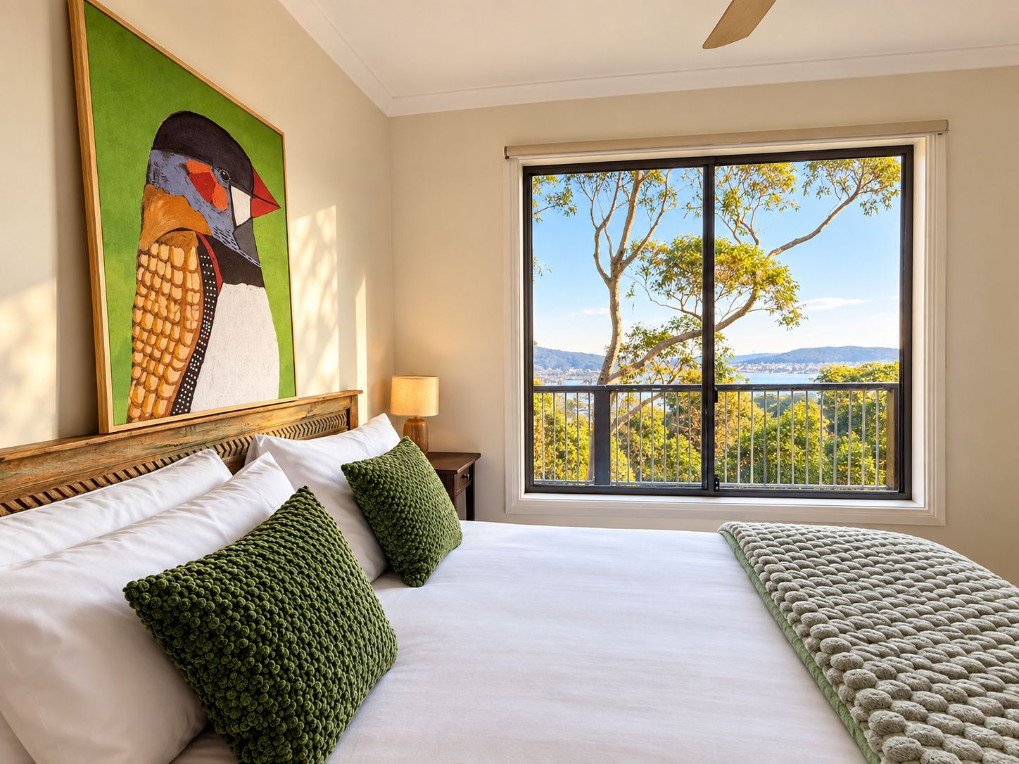 A hotel room with a large bed, a painting of a bird, a view of trees and mountains through a window, and a nightstand with a lamp.