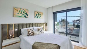 Quarto de hotel moderno com uma cama grande, duas obras de arte nas paredes, vista para a praia e uma varanda com cadeiras.