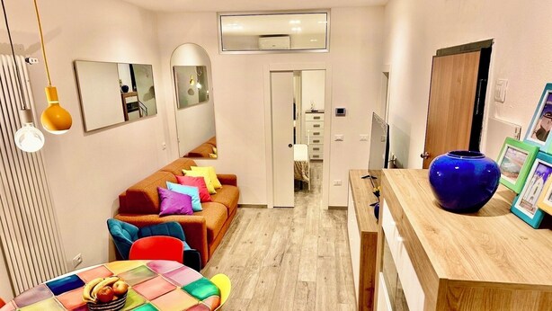 Ein modernes Wohnzimmer mit einer Couch, bunten Kissen, einem Couchtisch mit Früchten und einer blauen Vase auf einem Holznachttisch.