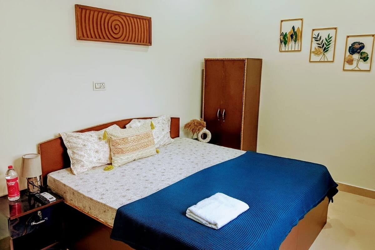 A neatly made bed with a patterned bedspread and pillows, a wooden wardrobe, a small bedside table with a lamp and a bottle, and a wall with three framed botanical prints.