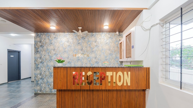 A reception area with a wooden counter, a ceiling fan, and a window with blinds. - Hotel Royal Manyaa (Bengaluru)