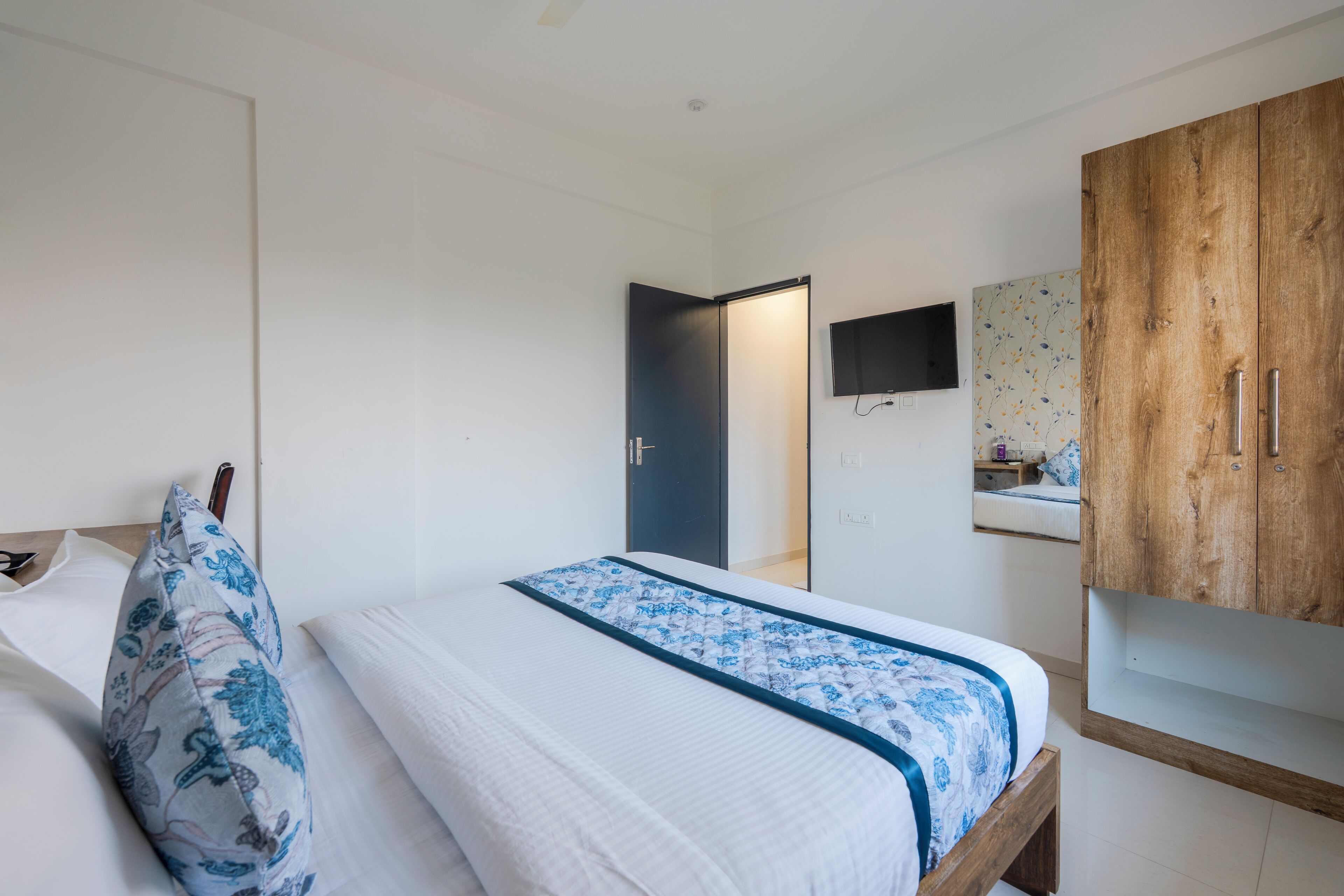 A bedroom with a bed, a wooden wardrobe, a TV mounted on the wall, and a bathroom with a sink and mirror.