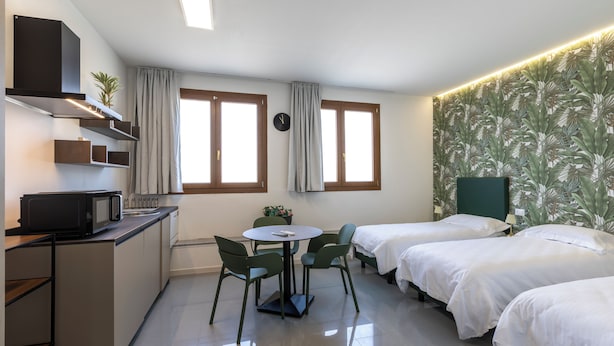 Quarto de hotel compacto com cozinha compacta, duas camas de solteiro, uma mesa de jantar redonda com cadeiras e uma janela grande com cortinas.
