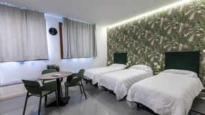 Quarto de hotel com duas camas, uma mesa redonda com cadeiras, um relógio na parede e um papel de parede tropical.