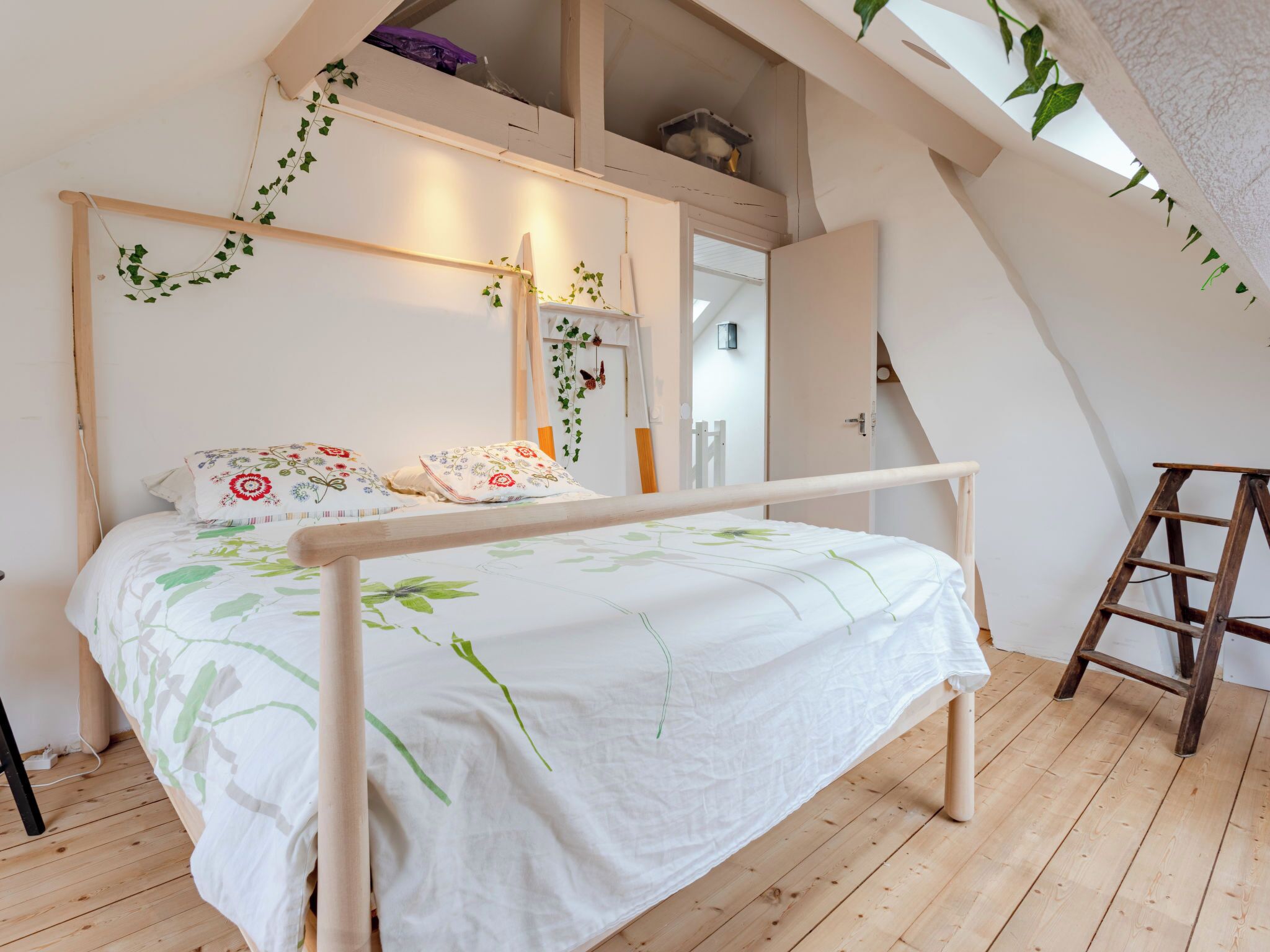 A bedroom with a wooden bed, a ladder, and a sloped ceiling with plants.