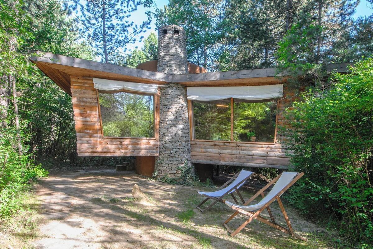 Un chalet rustique doté d’une cheminée en pierre, de grandes fenêtres et d’une terrasse en bois.